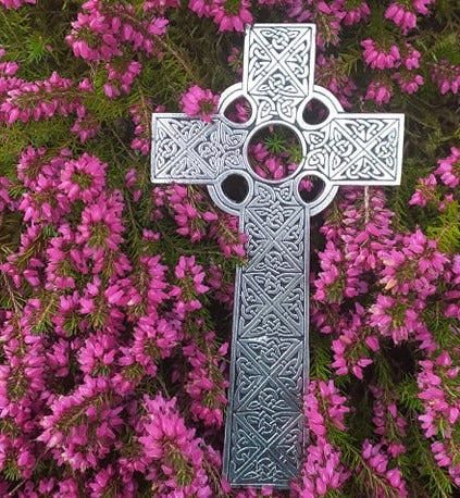 A handcrafted Mullingar Pewter Cross wall hanging with intricicate celtic knot work and resting on a bed of bright pink heather.