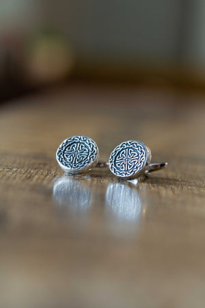 A pair of round silver-toned tie pins with an intricate Celtic knot design, resting on a wooden surface with a soft reflection.