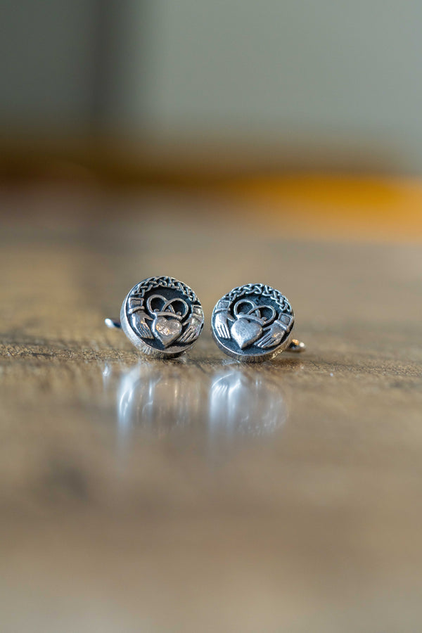 A pair of silver-toned tie pins with a Celtic claddagh design, resting on a reflective wooden surface with a blurred background.