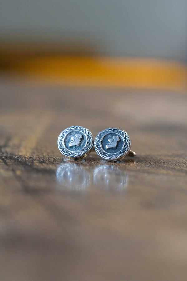 A pair of round silver-toned tie pins with a textured border and a central raised emblem, resting on a wooden surface with a soft reflection.