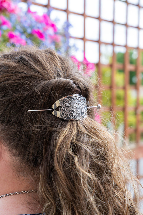 A close-up lifestyle shot of a woman's brown hair styled in a half-up bun, secured with an ornate silver metal hair claw clip. The background is a soft-focus outdoor scene with a wooden trellis and pink flowers.