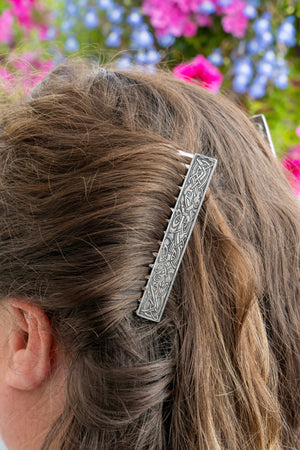 A close-up of a woman's hair secured with a decorative silver hair claw clip, with a blurred background of pink and blue flowers.