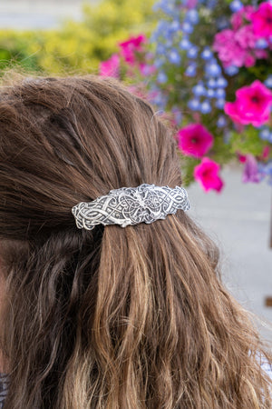 A close-up lifestyle shot of a person's brown hair tied back with a silver-toned, intricately woven hair accessory, set against a blurred background of colorful flowers.