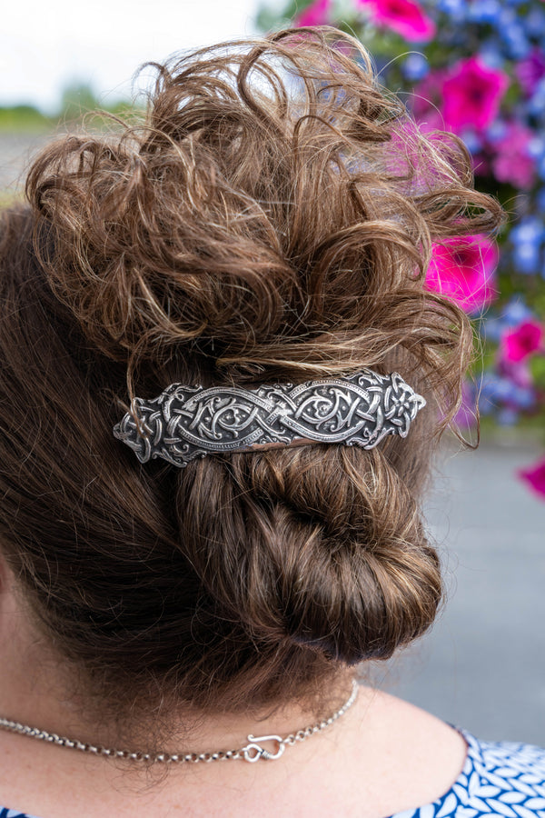 A close-up back view of a person wearing a silver-toned Celtic knot hair clip to hold up a messy bun, with blurred pink and blue flowers in the background.