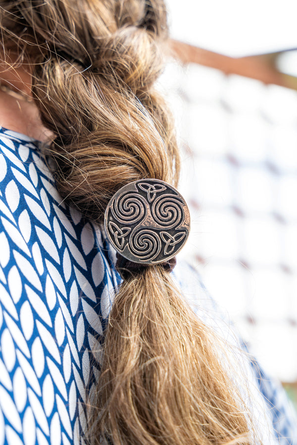 A close-up of a person's long light brown hair tied back with a round dark metal hair accessory featuring a Celtic knot swirl pattern, wearing a blue and white patterned top.