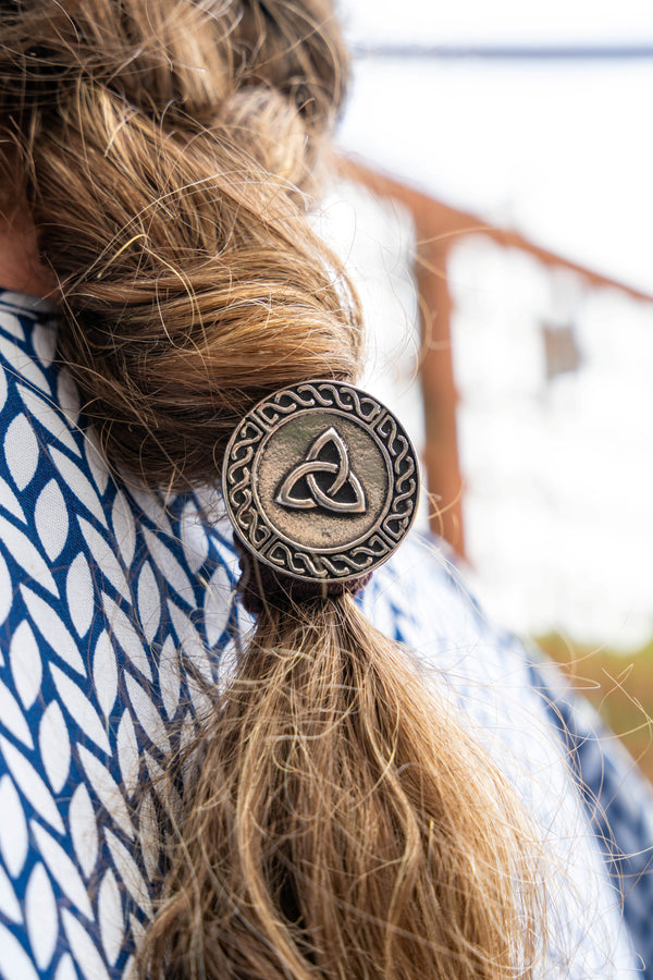 A close-up lifestyle shot of a person's brown hair tied back in a low braid with a round, antique bronze Celtic triquetra knot hair accessory.