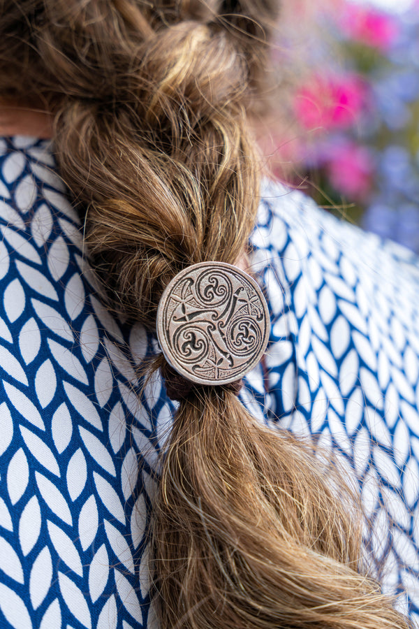 A close-up of a person's brown braided hair secured with a round silver-toned metal hair pin featuring an ornate Celtic knot design, wearing a blue and white patterned top.