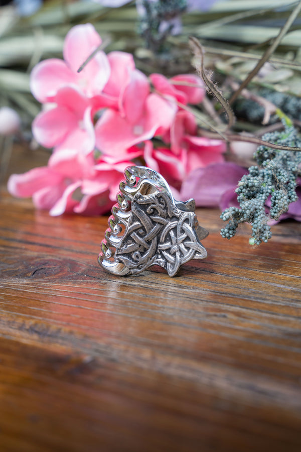 A silver-toned Celtic knot ring with a stylized animal head design, resting on a rustic wooden surface with pink flowers in the blurred background.