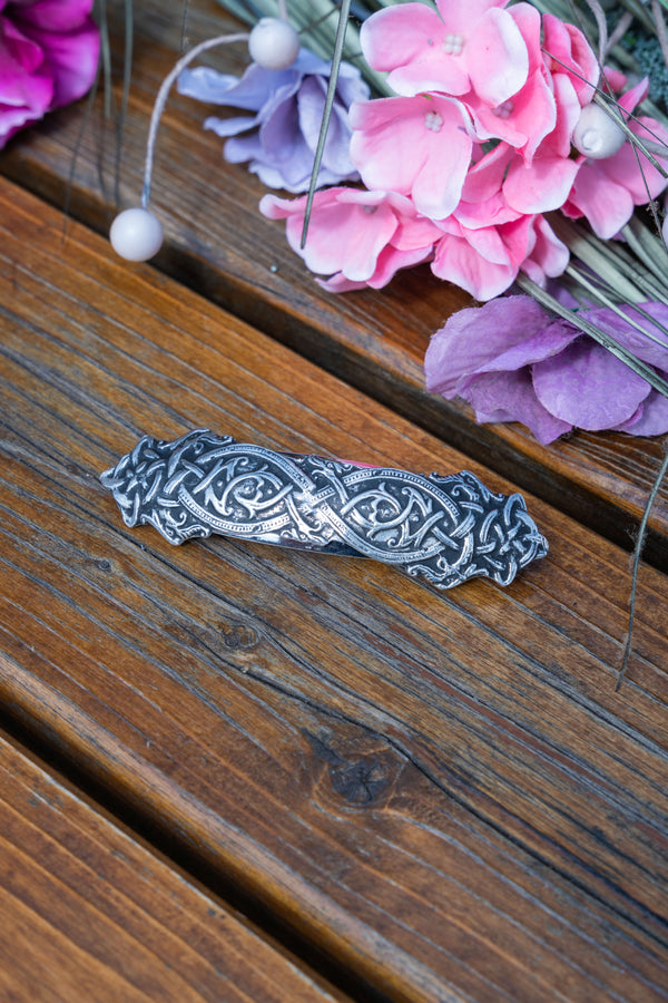 A silver-toned metal bar with an intricate Celtic knot pattern, resting on a rustic wooden surface with pink and purple flowers in the background.