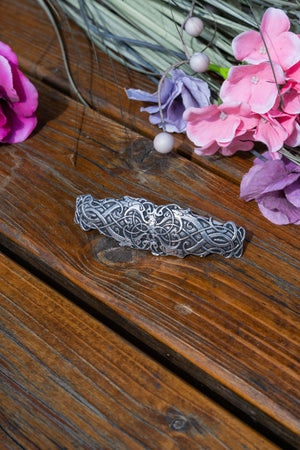 A silver-toned Celtic knotwork hair claw clip resting on a rustic wooden surface, styled with pink and purple flowers in the background.