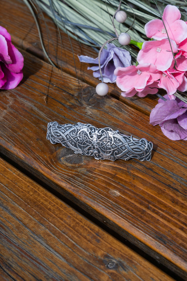 A silver-toned Celtic knotwork hair claw clip resting on a rustic wooden surface, styled with pink and purple flowers in the background.