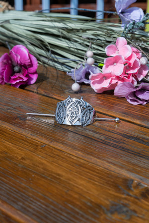 A silver-toned metal hair pin with an intricate Celtic knot design, displayed on a rustic wooden surface with pink and purple flowers in the background.
