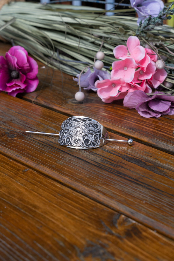 A silver-toned ornate brooch with a pinback, displayed on a rustic wooden surface with pink and purple flowers in the background.