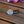 Load image into Gallery viewer, A round silver-toned metal hair tie cap with an engraved Celtic knot pattern, attached to a black elastic band, resting on a weathered wooden surface with pink and purple flowers in the background.
