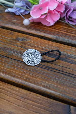 A round silver-toned metal hair tie cap with an engraved Celtic knot pattern, attached to a black elastic band, resting on a weathered wooden surface with pink and purple flowers in the background.