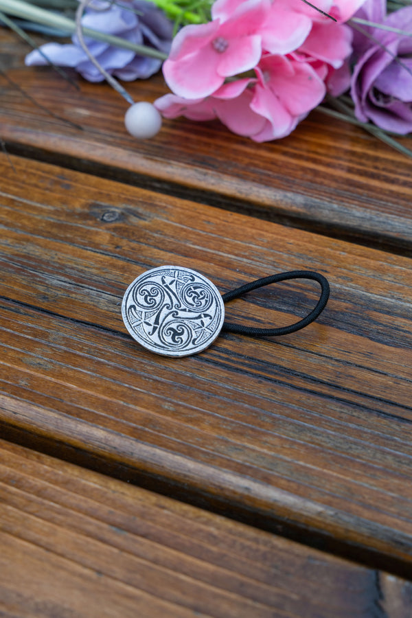 A round silver-toned metal hair tie cap with an engraved Celtic knot pattern, attached to a black elastic band, resting on a weathered wooden surface with pink and purple flowers in the background.