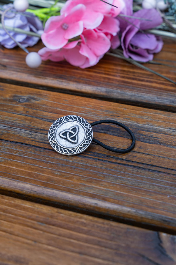 A silver-toned Celtic triquetra knot hair tie with a black elastic band, resting on a rustic wooden surface with pink and purple flowers in the background.