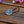 Load image into Gallery viewer, A black elastic hair tie with a round, silver-toned metal charm featuring an embossed Celtic cross, resting on a rustic wooden surface with blurred pink and purple flowers in the background.
