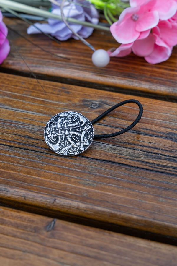 A black elastic hair tie with a round, silver-toned metal charm featuring an embossed Celtic cross, resting on a rustic wooden surface with blurred pink and purple flowers in the background.