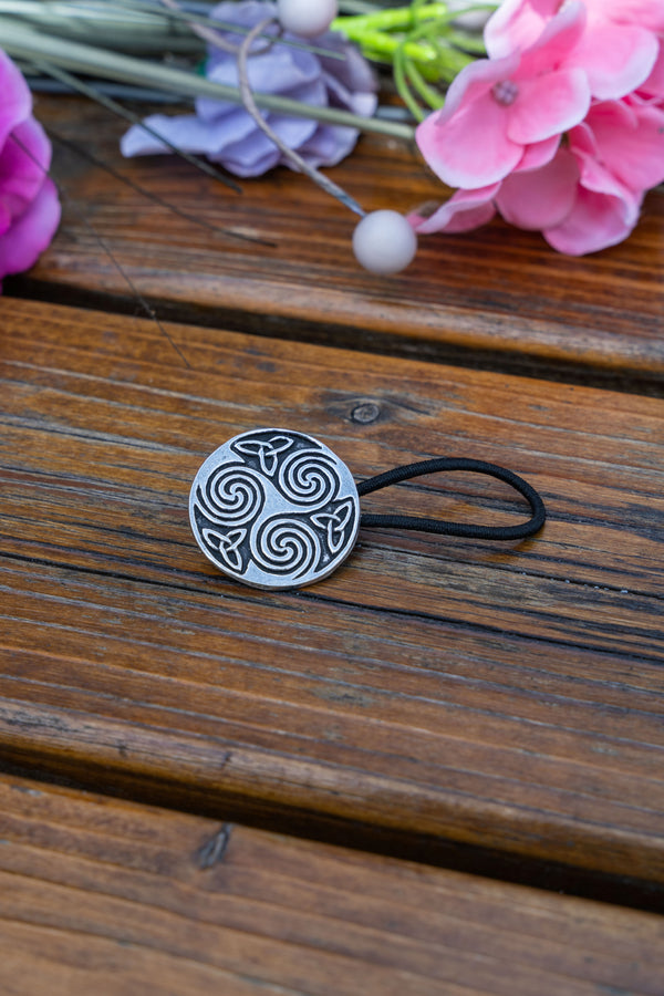 A silver Celtic knot circular hair tie clip with a black elastic band, resting on a rustic wooden surface with pink and purple flowers in the background.