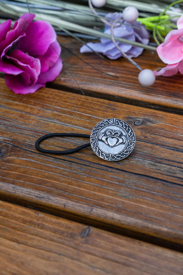 A silver-toned circular hair tie charm with an engraved heart design, attached to a black elastic band, displayed on a rustic wooden surface with decorative flowers.