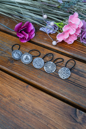 Four black elastic hair ties with silver Celtic knot and triquetra pendant charms, arranged on a rustic wooden surface with pink and purple flowers in the background.