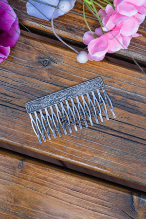 An ornate silver metal hair comb with engraved floral patterns on the bar, displayed on a rustic wooden surface with pink flowers in the corner.