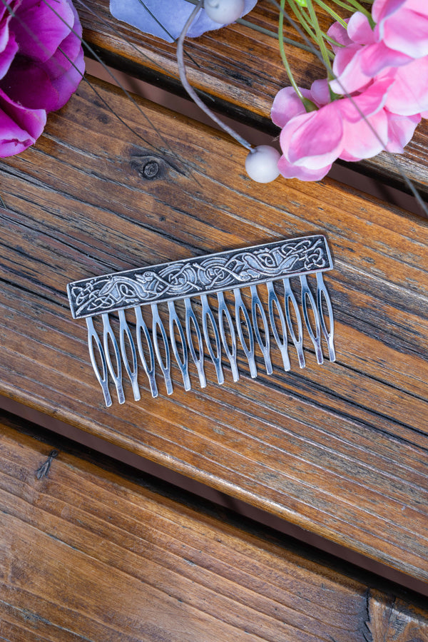 An ornate silver metal hair comb with intricate Celtic knotwork engraving on the rectangular head, displayed on a rustic wooden surface with pink flowers in the background.