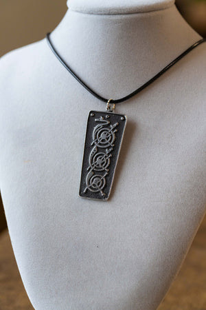 A rectangular black pendant necklace with an engraved circular symbol, displayed on a white fabric mannequin bust.