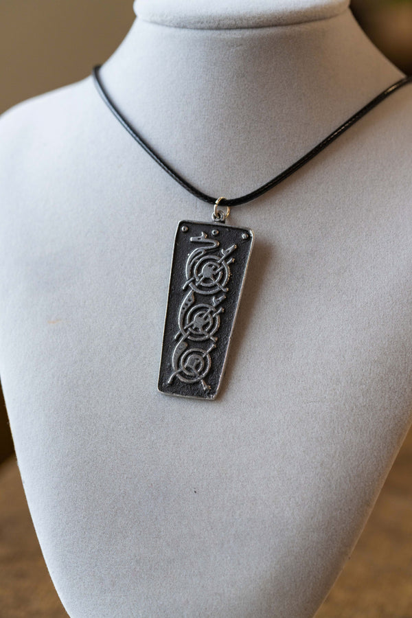 A rectangular black pendant necklace with an engraved circular symbol, displayed on a white fabric mannequin bust.