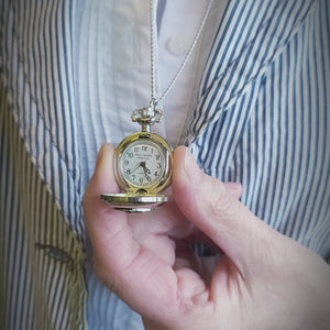A lady wearing a stripped Jacket and white blouse holding at chest height a Mullingar Pewter Necklace. The Pendent of which is a watch. 