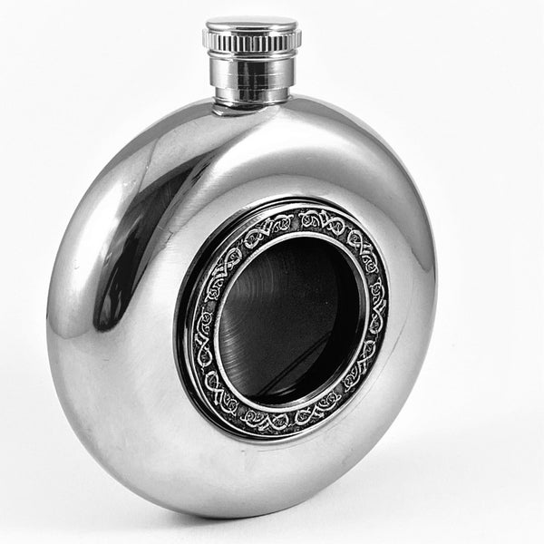 WHISKEY FLASK WITH GLASS CENTRE AND CELTIC KNOT DESIGN THAT SURROUNDS THE GLASS CENTER ARE A COMBINATION OF PEWTER AND STAINLESS STEEL IN A POLISHED SILVERWARE LOOK. THE FLASK HOLDS 5 FLUID OZS AND STANDS 4" TALL. MADE IN IRELAND BY MULLINGAR PEWTER